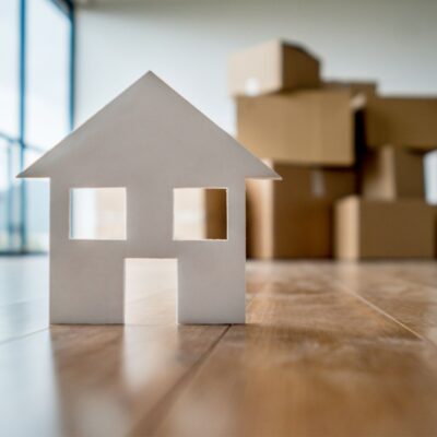 Debt-to-income for Travel Nurses. White paper house cutout in front of brown moving boxes.